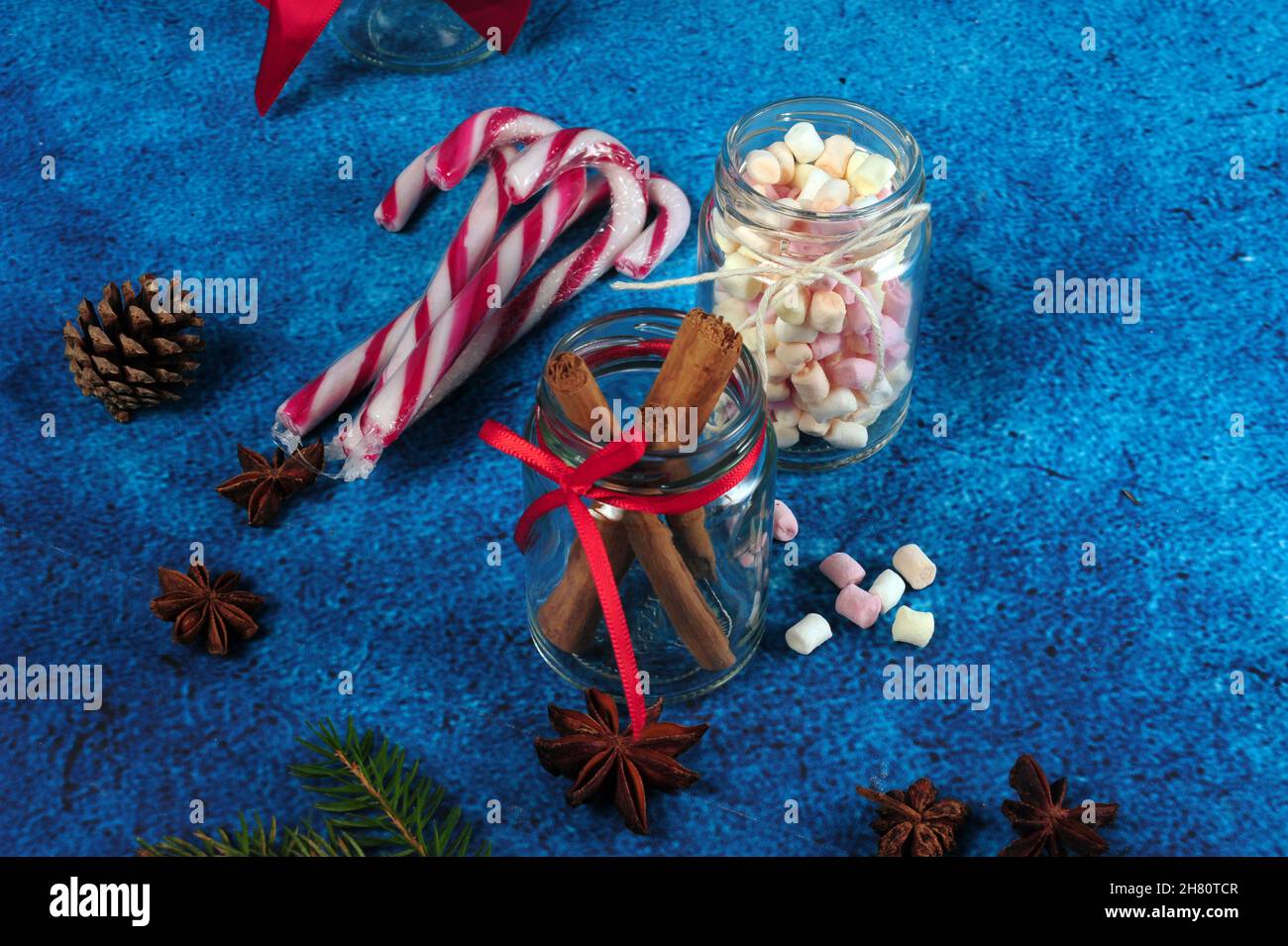 Jars filled with mini marshmallows and cinnamon sticks, and pinecone on a blue surface Stock Photo