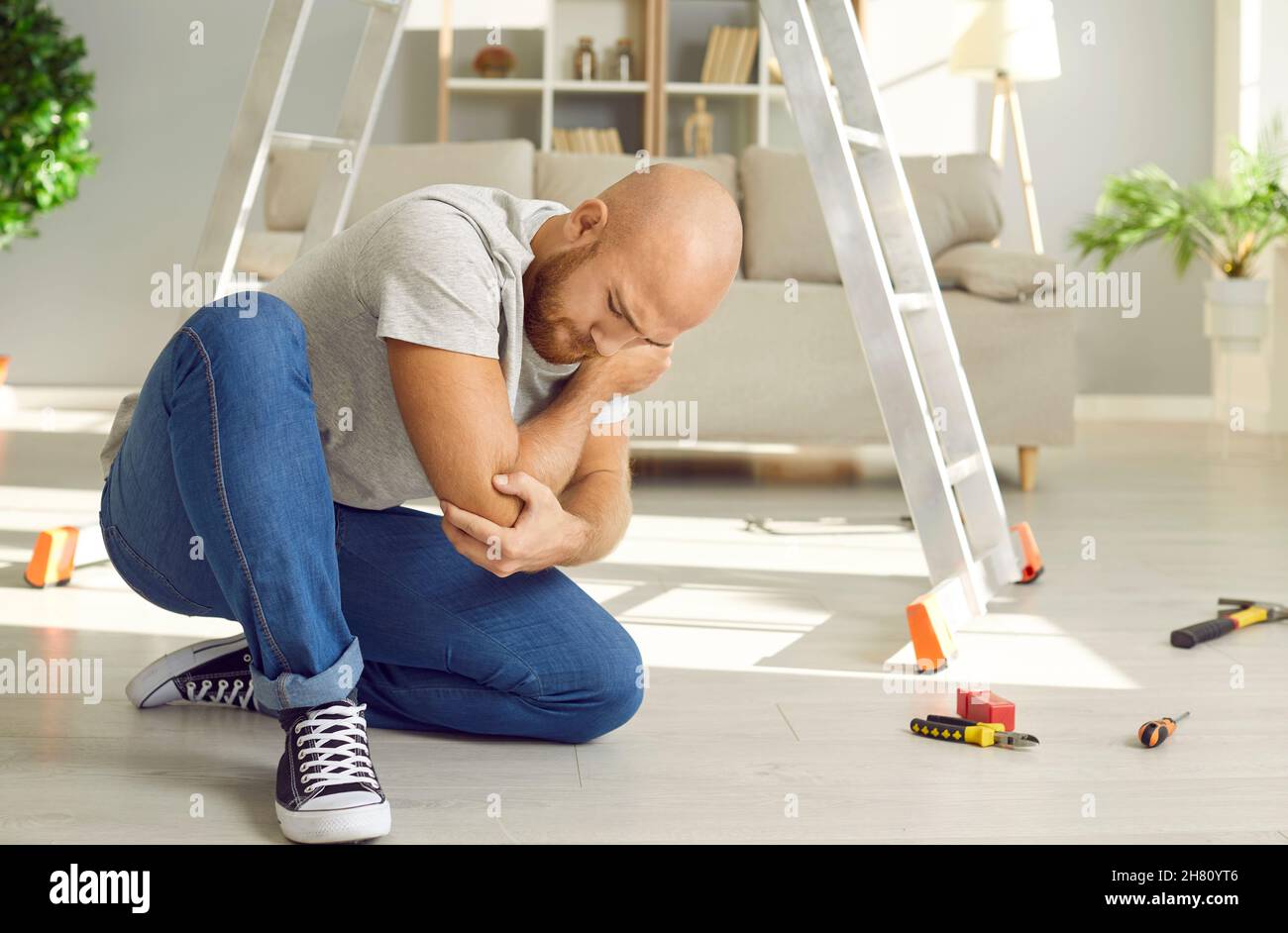 Man who just fell off ladder at home is sitting on floor and touching his hurt elbow Stock Photo