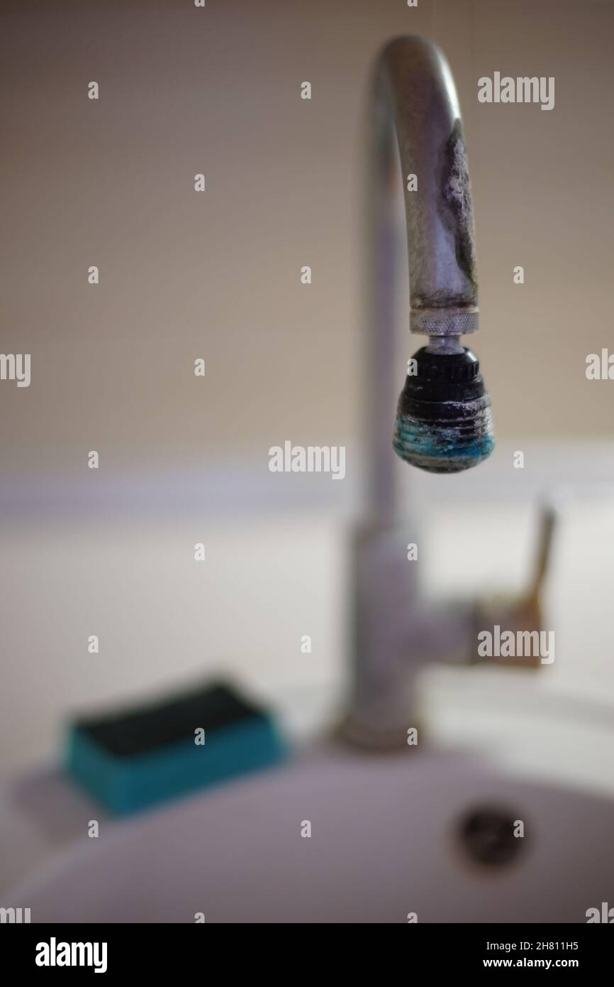 The old chrome faucet in the kitchen sink is covered in limescale. Blue dish sponge lies on a white sink. Stock Photo