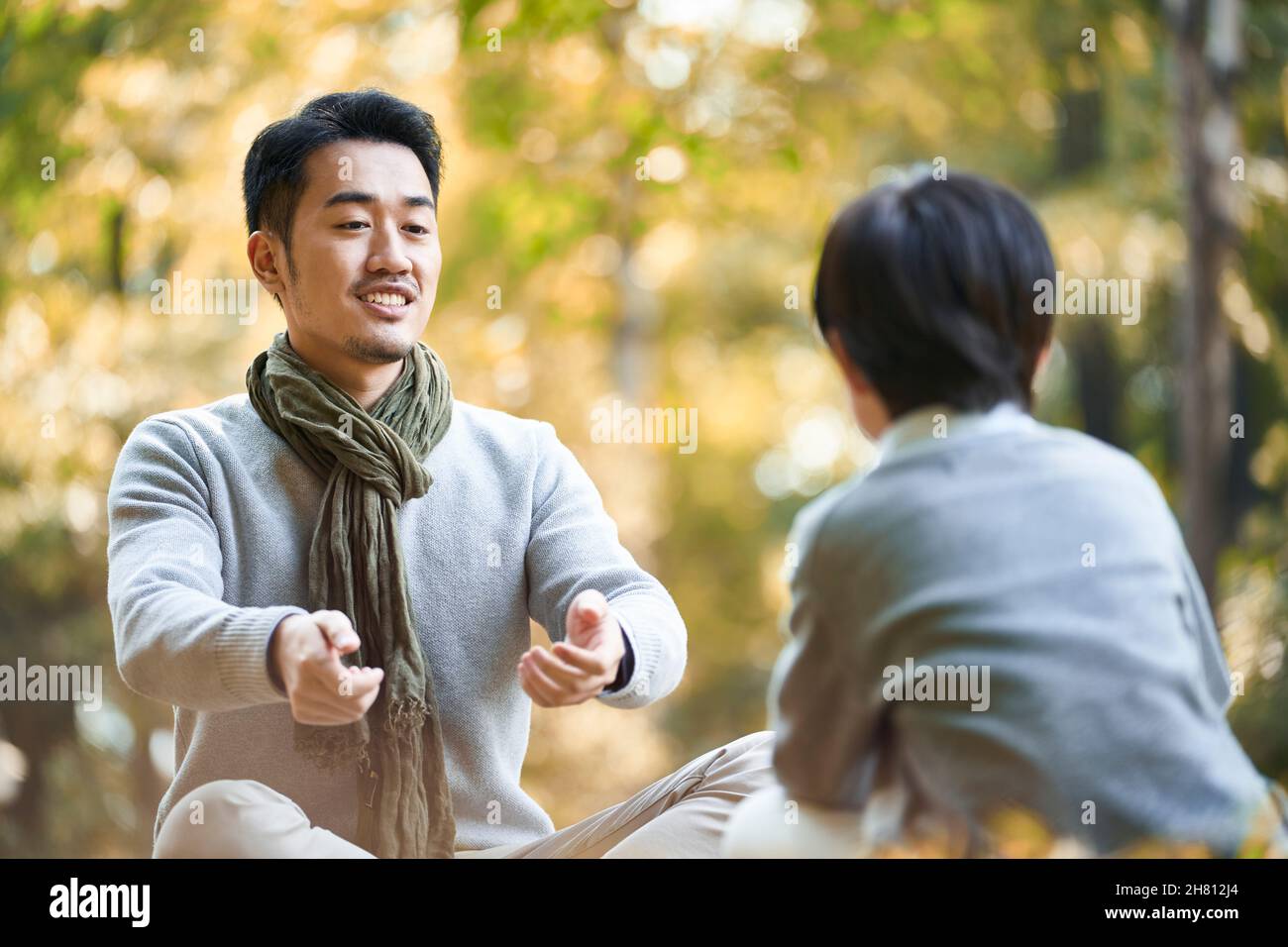 asian father and son sitting on grass having a pleasant conversation outdoors in park Stock Photo