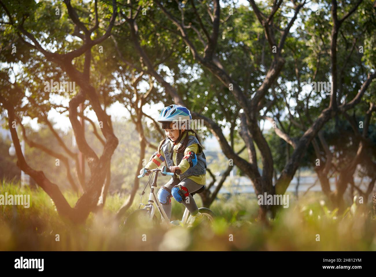 lovely happy asian little girl with helmet and full protection gear riding bike in city park Stock Photo