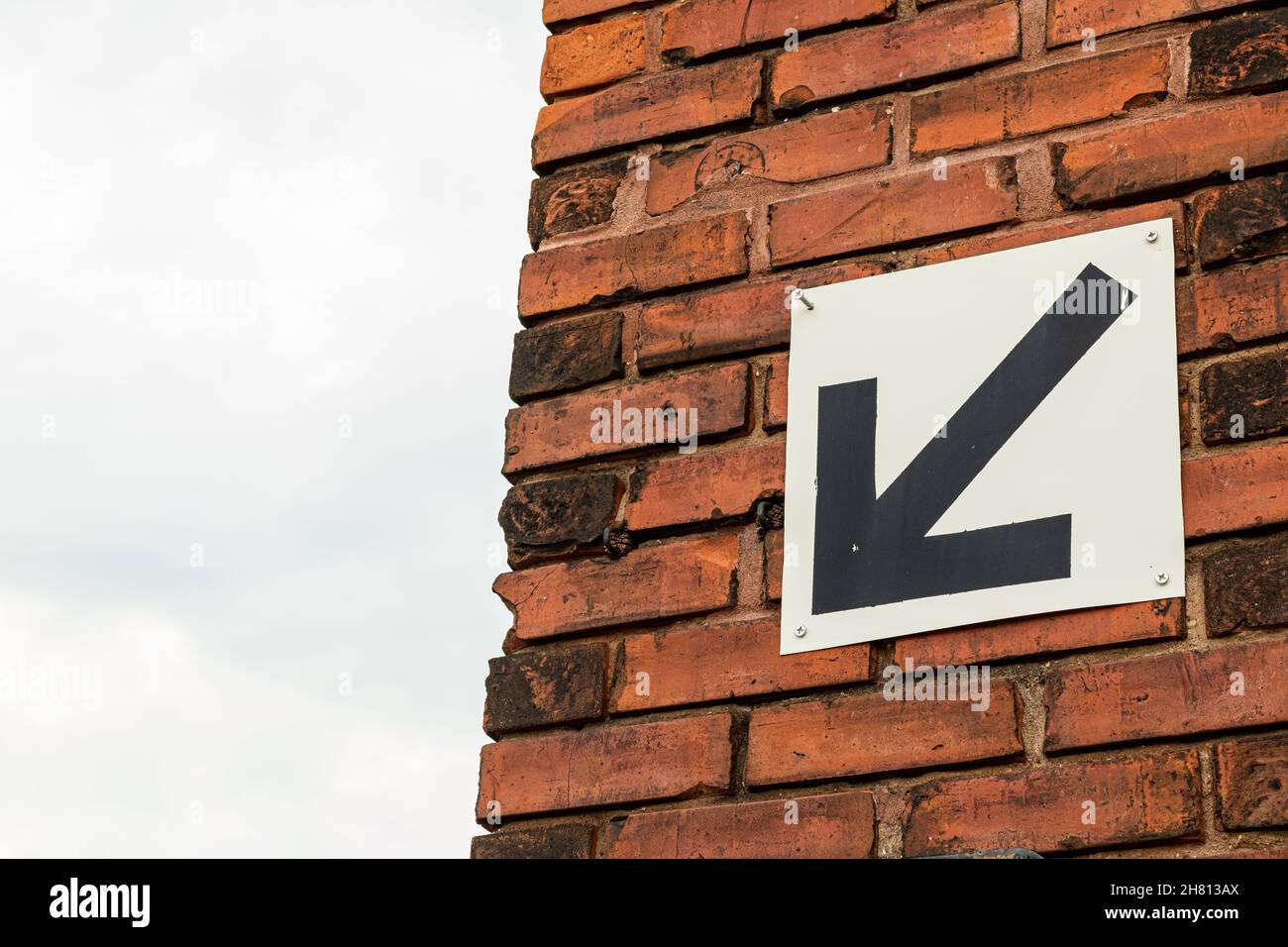 Black arrow on white background pointing down and left. Against background of red brick wall and blue sky. Copy space. Stock Photo