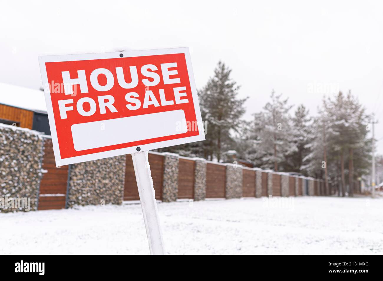 Red sign - house for sale, outdoors in winter against the backdrop of a fence and snowy pine trees. Buying suburban real estate with a mortgage. Sale Stock Photo