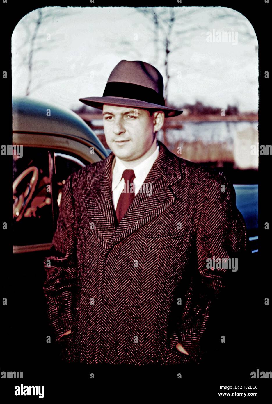 Vintage Kodachrome slide circa 1949 of a young man in his 20s dressed in tweed overcoat, hat, tie and white shirt by a 1940s automobile. Stock Photo