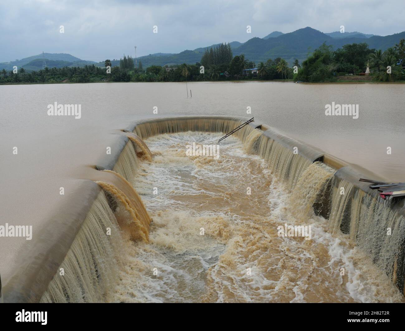 Brown water in the dam overflowing into the spillway with mountain and green forest in background , Flood in rainy season, Thailand Stock Photo