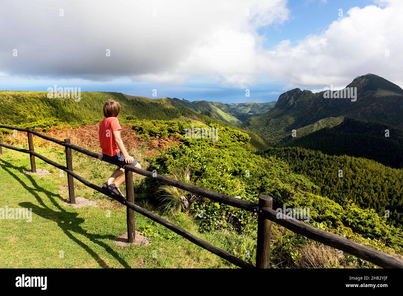 Boy in red t shirt sitting on wooden fence at viewpoint Miradouro do Vale da Fazenda viewpoint in Flores, Azores, Portugal Stock Photo