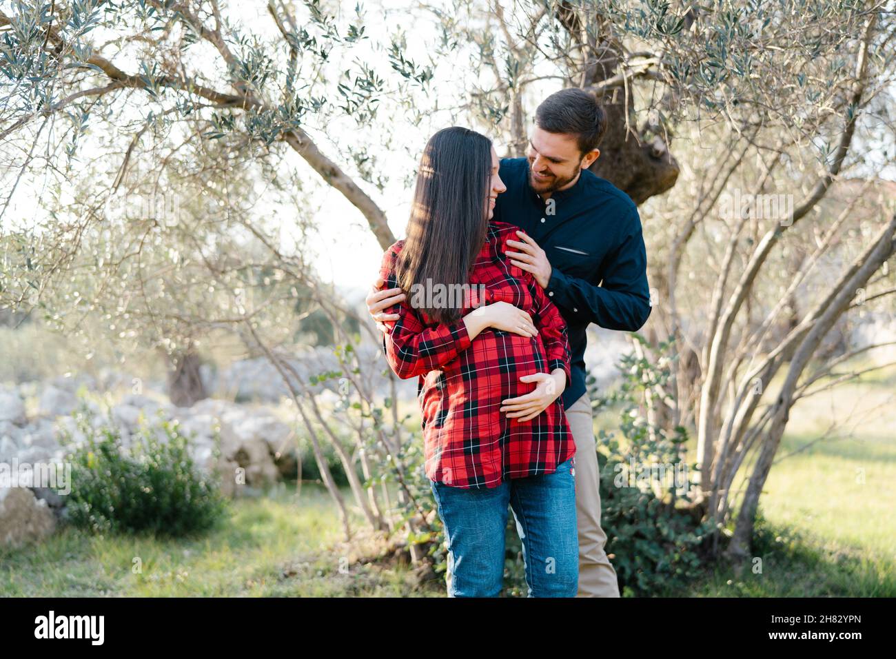 Smiling man hugging shoulders of pregnant woman from behind in park ...