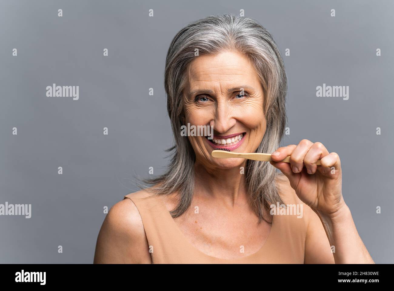 Headshot of smiling senior gray-haired woman with bamboo toothbrush brushing her white teeth isolated on gray background, everyday care routine. Dental care and oral hygiene Stock Photo