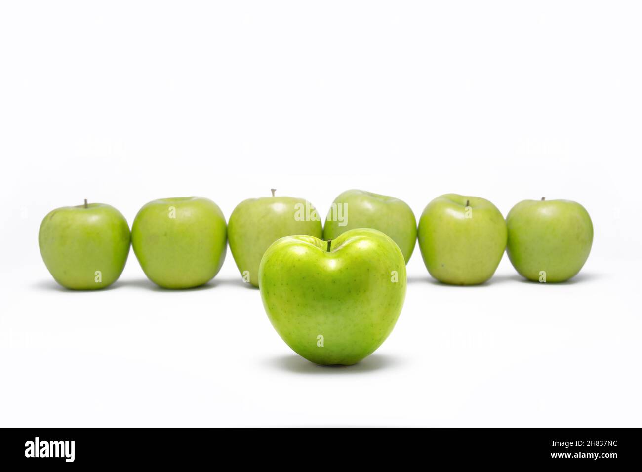 Green apple heart shaped in front of other green apples in line. On white background. Stock Photo
