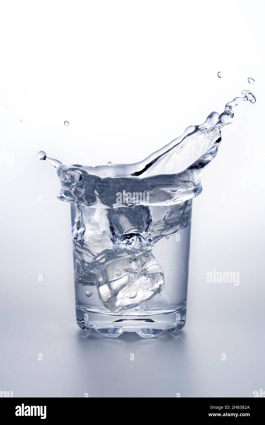 Glass of water with ice cube splashing. On grey surface and white background. Stock Photo
