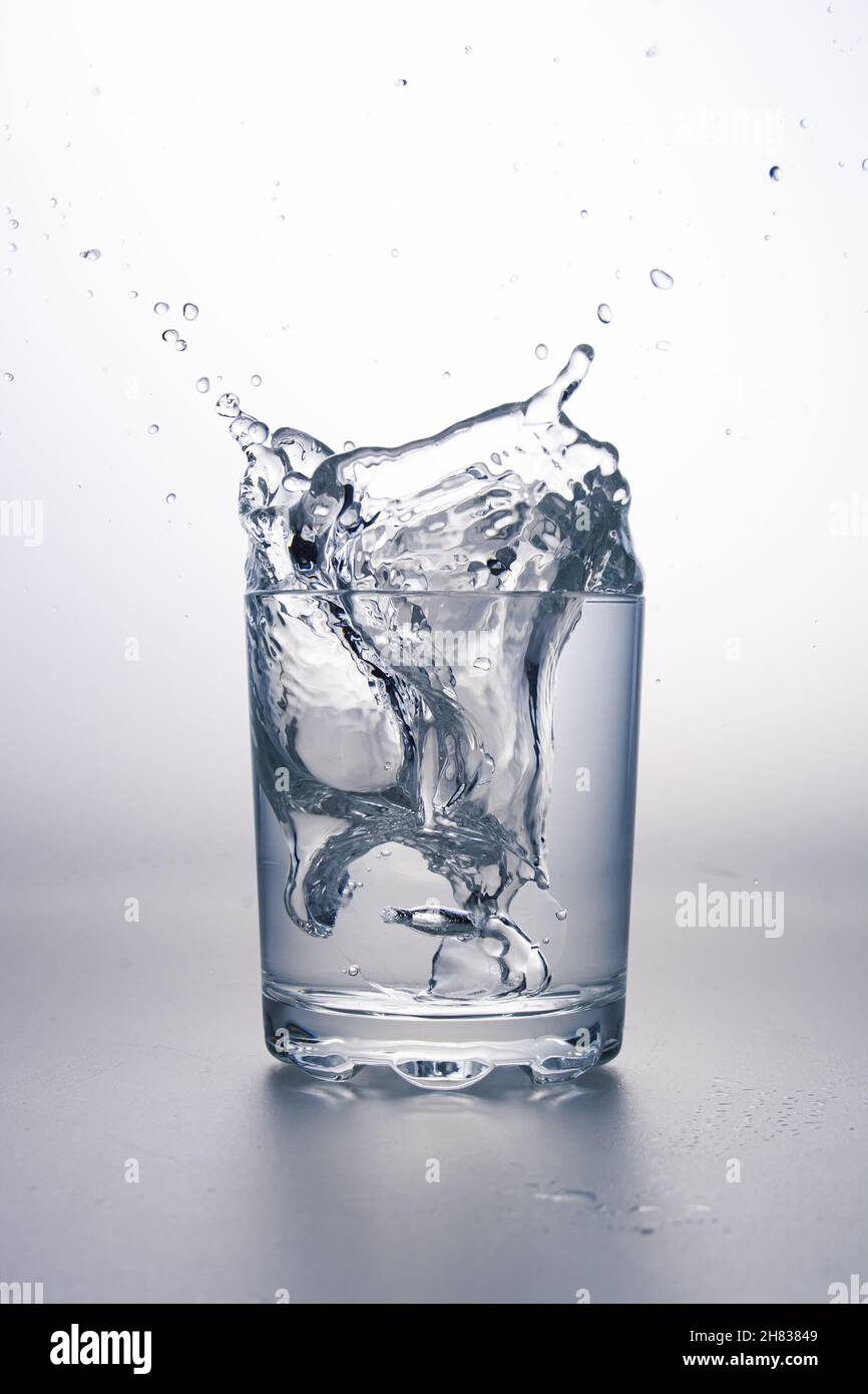 Glass of water with splash on light grey surface and white background. Stock Photo