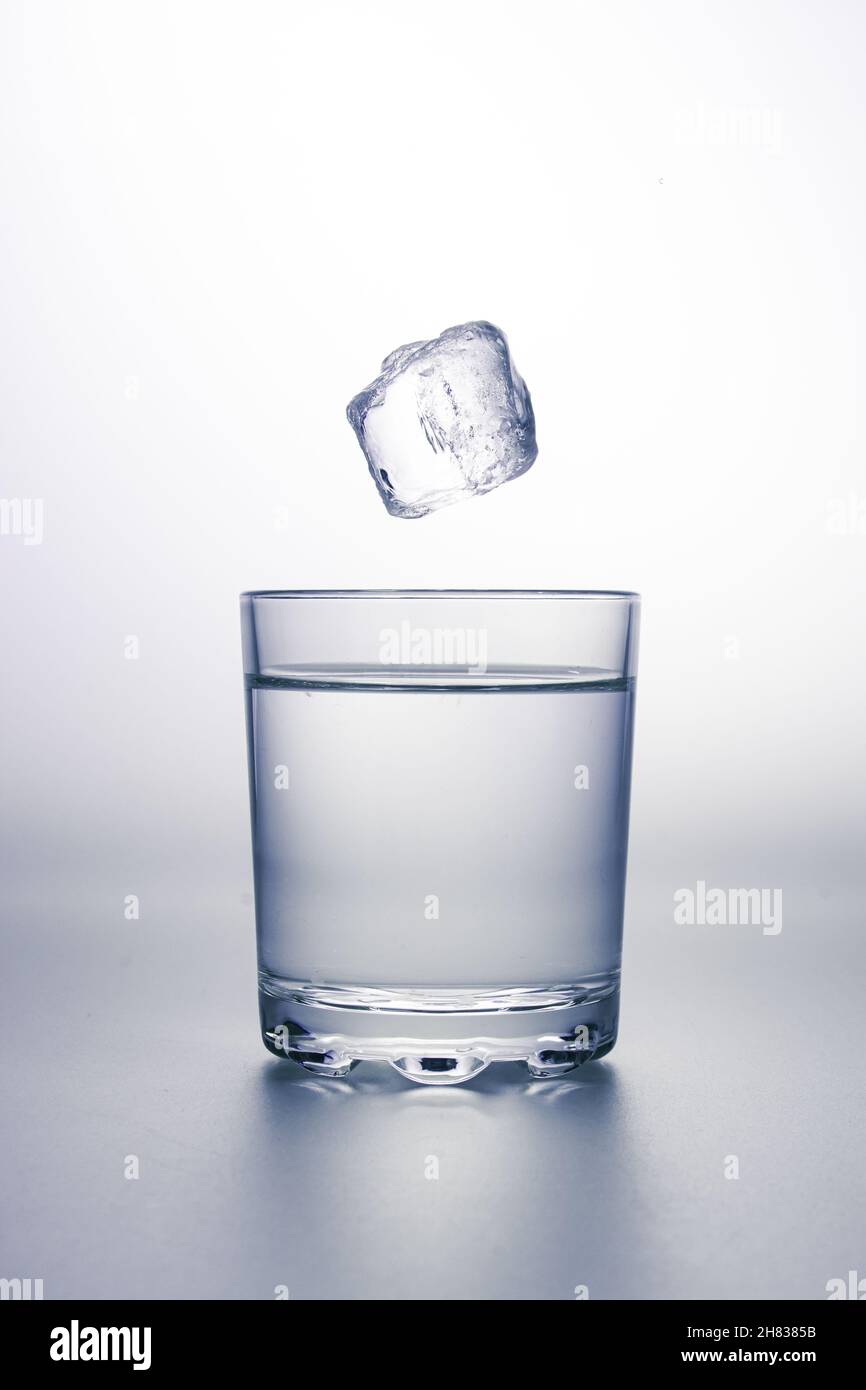 Glass of clear water with an ice cube falling above. On grey surface and white background. Stock Photo