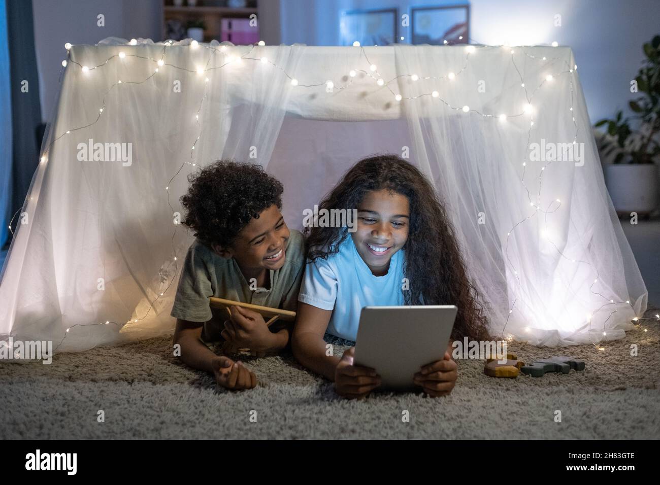 Cheerful African American brother and sister lying on carpet behind table covered with fabric and lights and using tablet Stock Photo