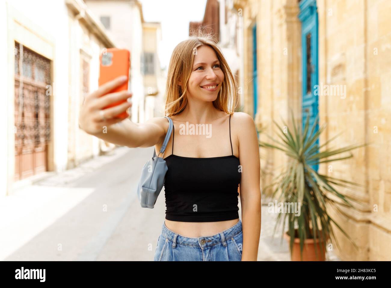 Young beautiful girl in denim jeans and purse handbag makes selfie against the background of the Cyprus old town. Stock Photo