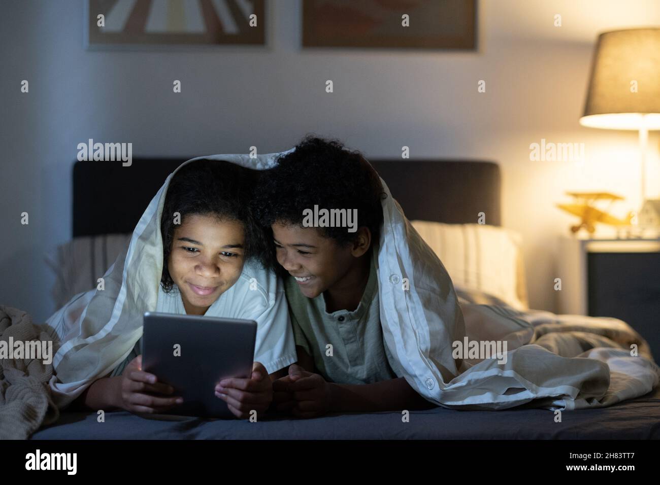 Positive black siblings lying under duvet on bed and using tablet while browsing internet at night Stock Photo