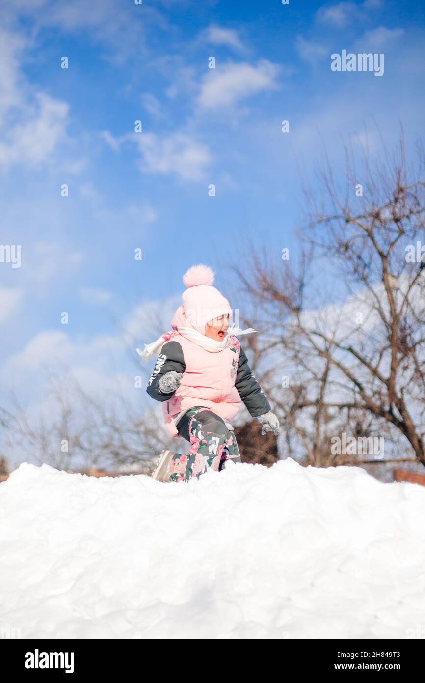 Child rides on snow slide. Little girl in winter warm suit plays in fresh air and enjoys sunny day in winter, she lies in snow and plays snowballs Stock Photo