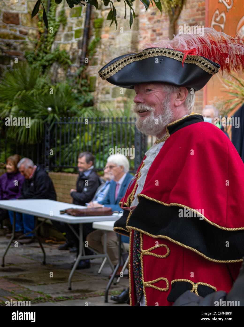 A man dressed as a town crier in the uk, wearing a red cloak with black and gold trim and a black tricorn hat with a peacock feather. White beard. Stock Photo