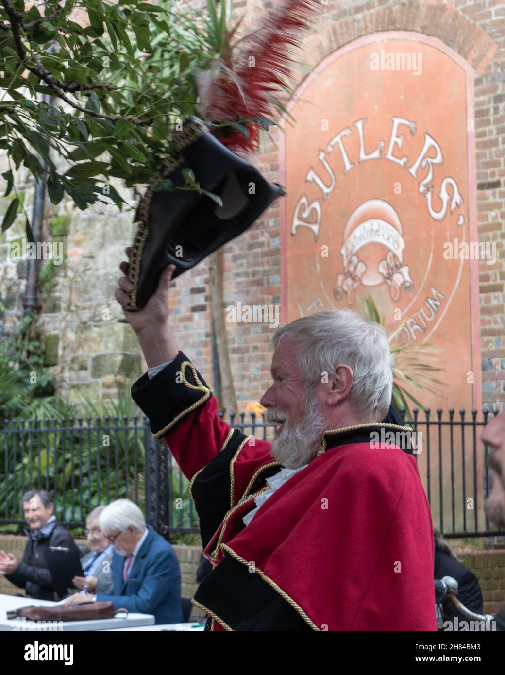 A man dressed as a town crier in the uk, wearing a red cloak with black and gold trim and a black tricorn hat with a peacock feather. White beard. Stock Photo