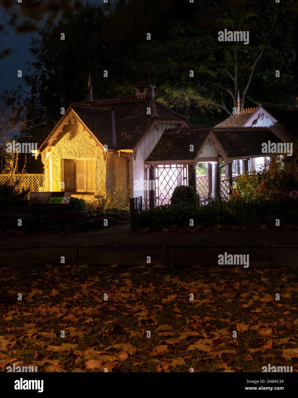 A night shot of Duck Island Cottage, St James's park, illuminated by warm yellow light with red/orange plane tree leaves on the ground. Stock Photo