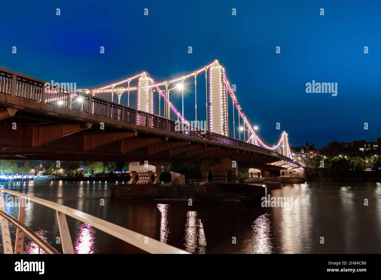A night times shot of Chelsea Bridge taken from the south bank of the river thames which shows the bridge lit up in warm white lights. River Thames Stock Photo