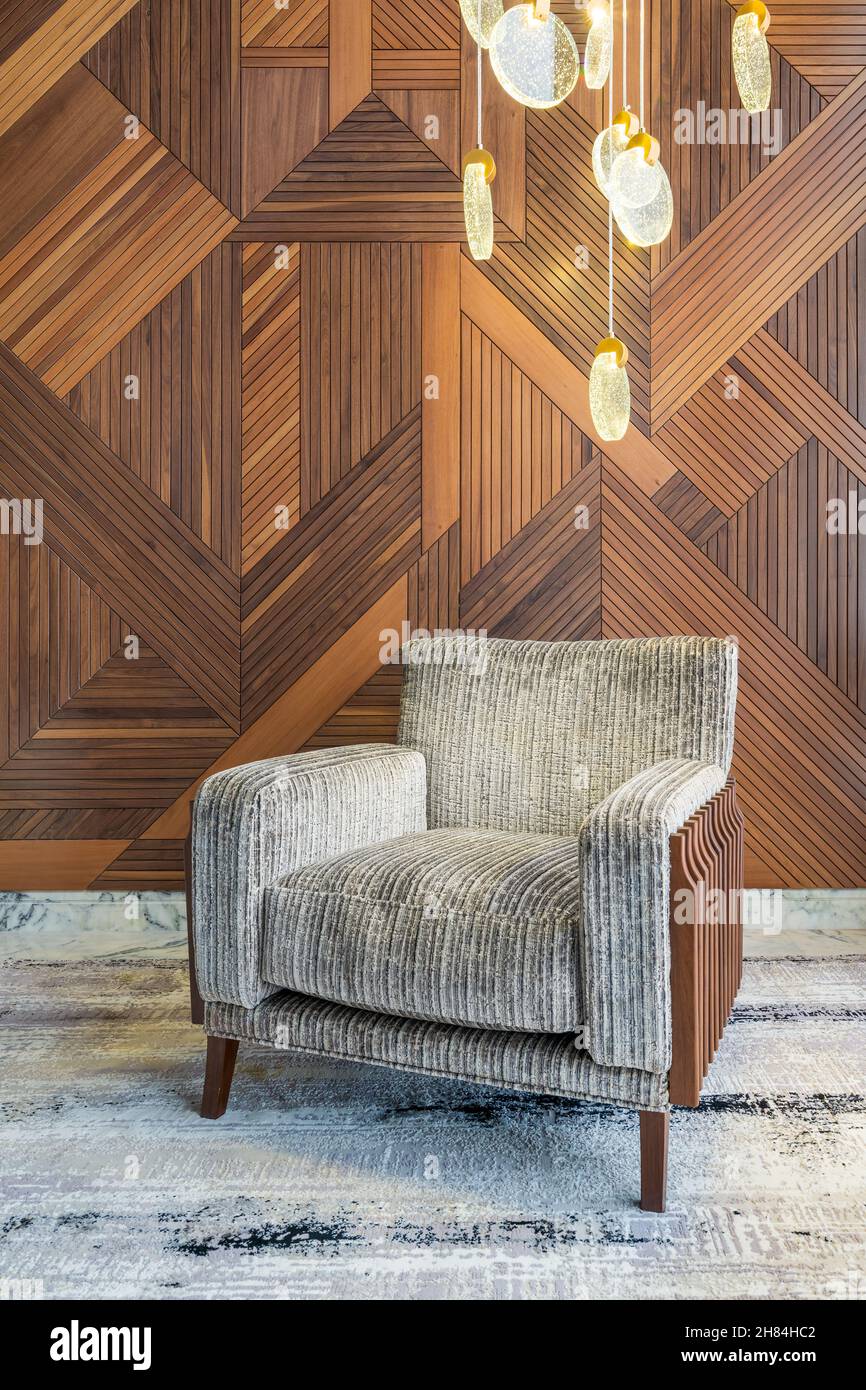 Modern grey armchair, and contemporary tall glass chandelier, in a hall with decorated wood cladding wall, and grey modern carpet Stock Photo