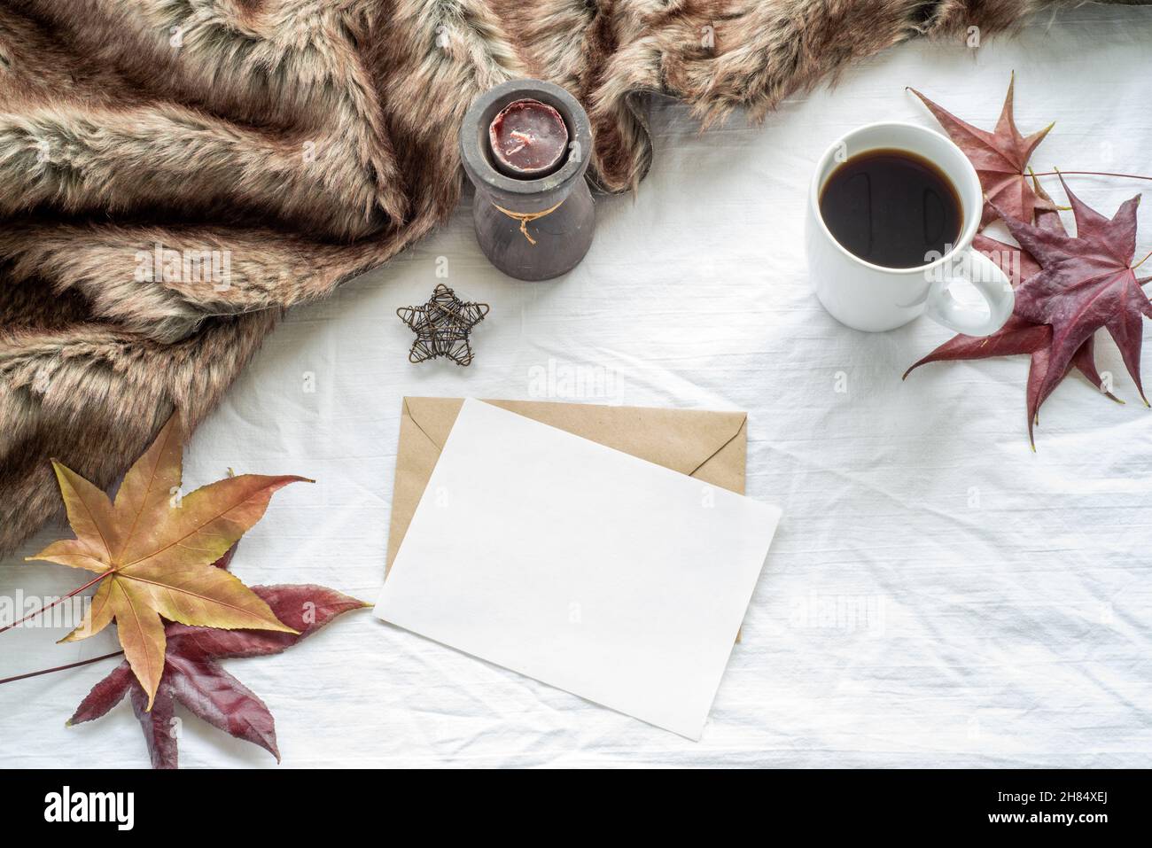 Autumn breakfast scene. Blank greeting card, invitation card mockup with maple leaves, cup of coffee, scented candle and plaid. White tablecloth backg Stock Photo