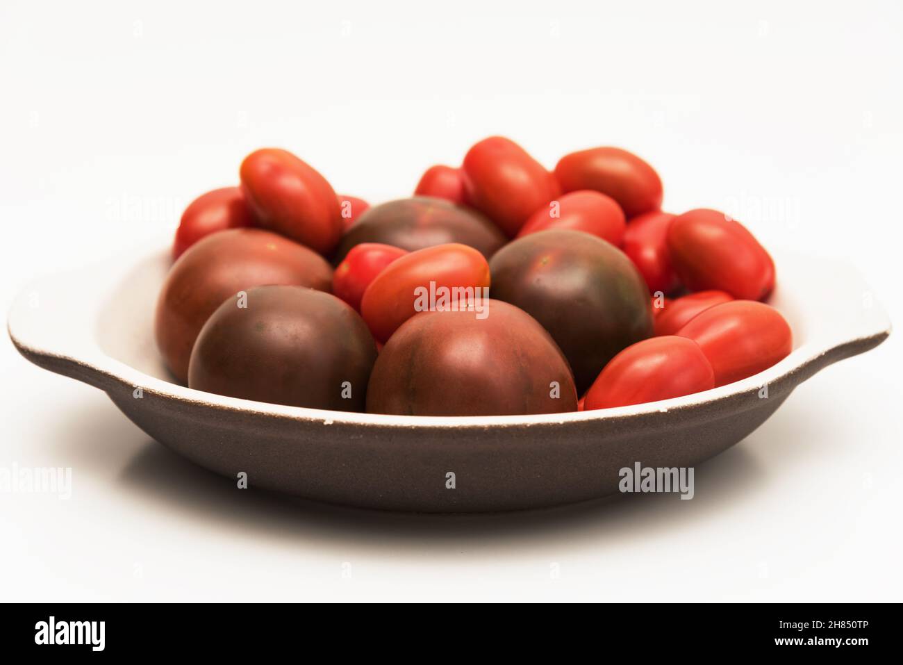 Delicious kumato and cherry tomatoes piled on a clay plate over white