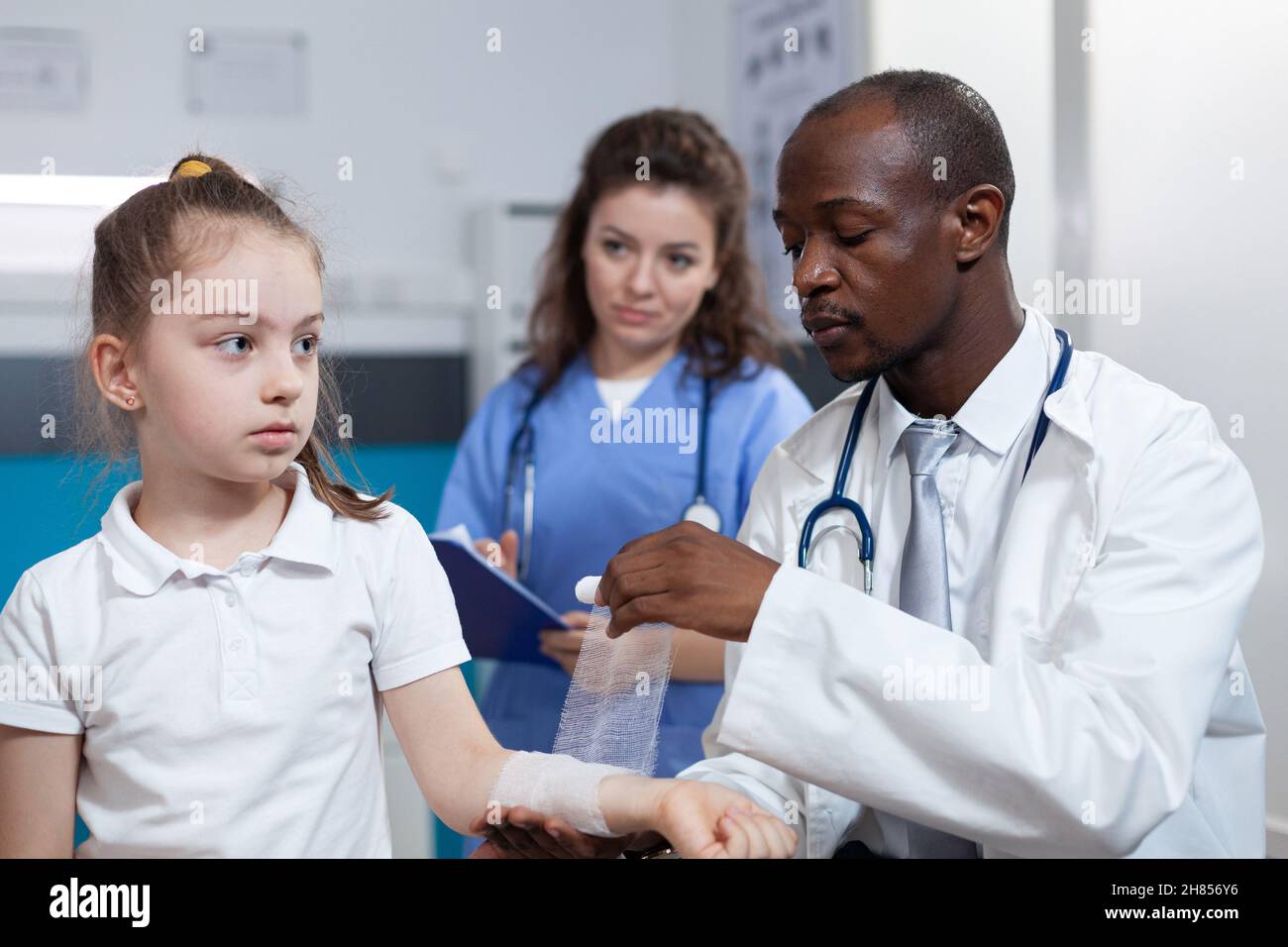 African american pediatrician doctor bandage fractured arm of girl patient during clinical inspection in hospital office. Young child having broken hand after accident. Health care service Stock Photo