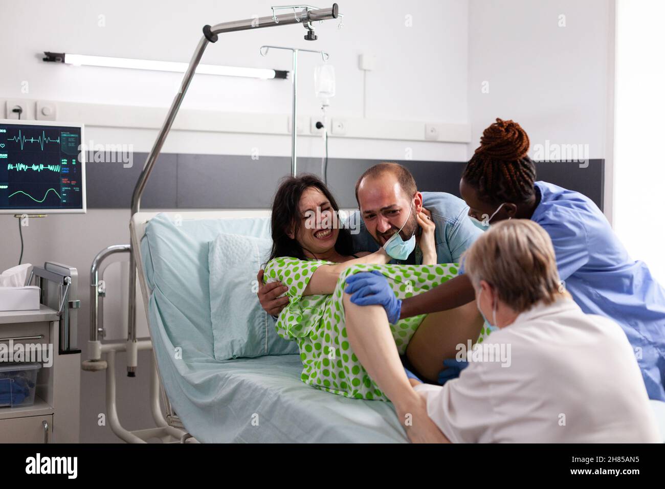 Young woman giving birth to child in hospital ward maternity with assistance from doctor and african american nurse. Multi ethnic medical team helping pregnant adult with baby delivery Stock Photo