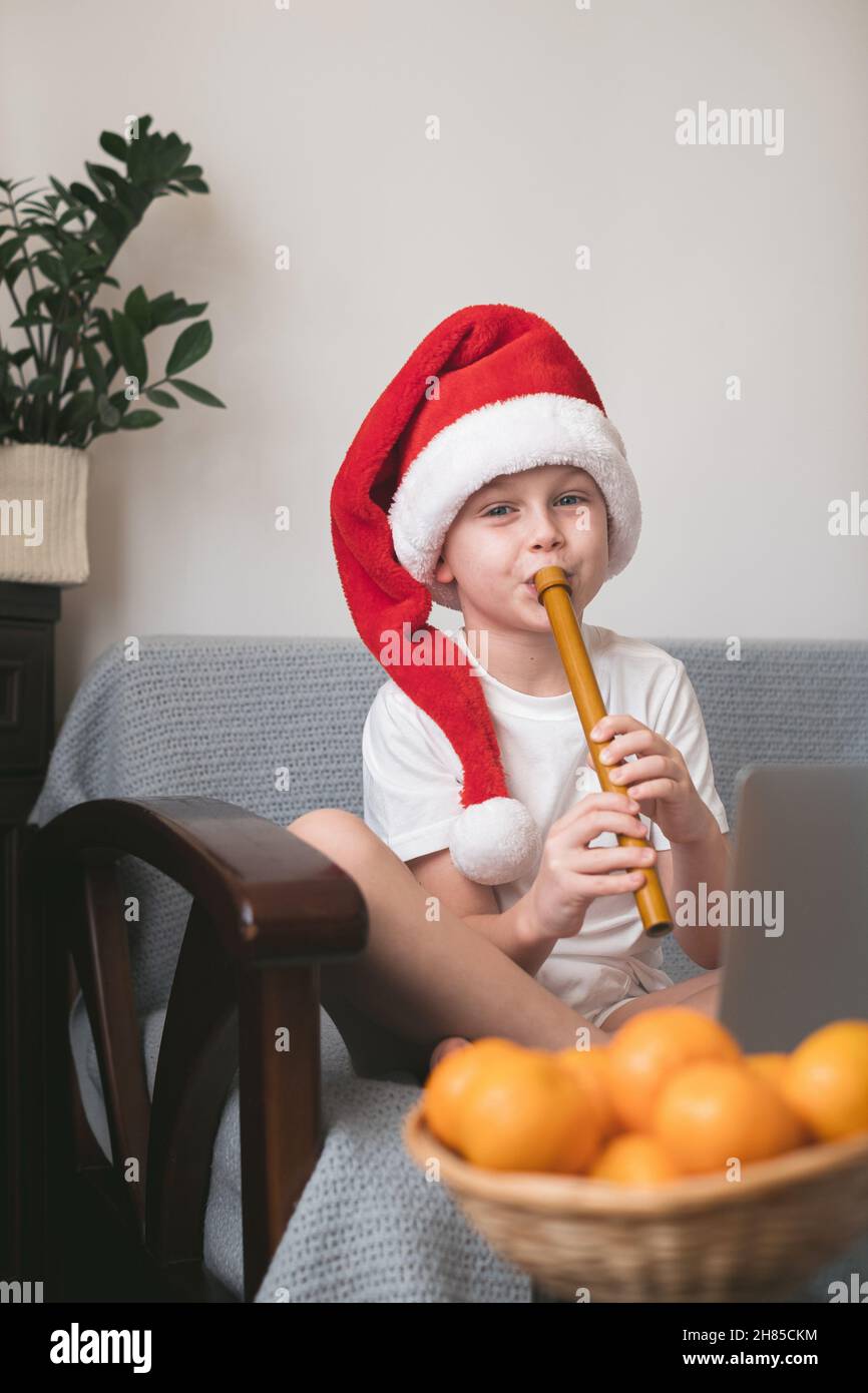 Boy in Santa hat learning to play the flute watching online course on laptop while practicing at home. Online training, online classes. Stock Photo