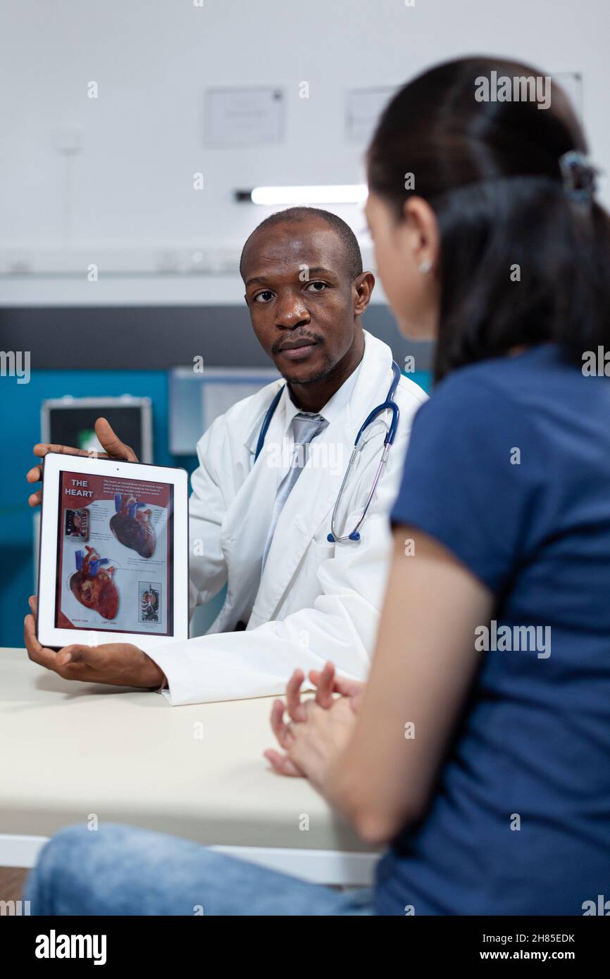 African american pediatrician doctor holding tablet computer in hands with heart radiography on screen discussing medical treatment with patient mother in hospital office. Health care service Stock Photo