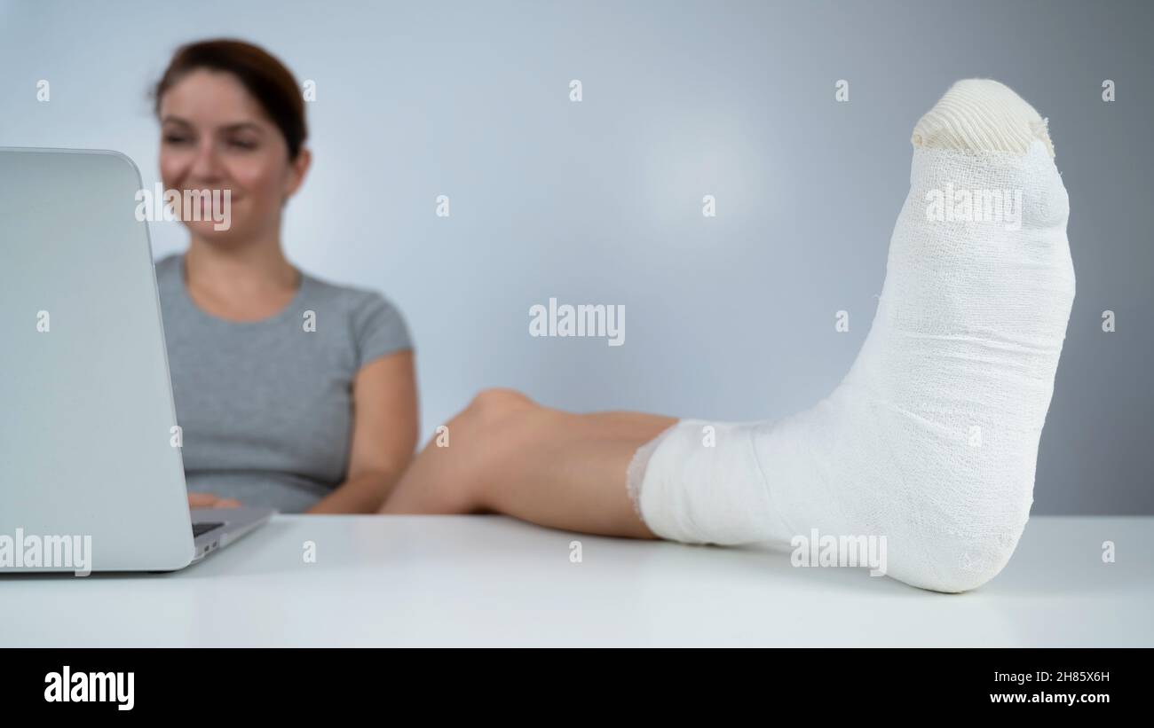 Caucasian woman lifted her leg with plaster to work desk and works on laptop on white background Stock Photo