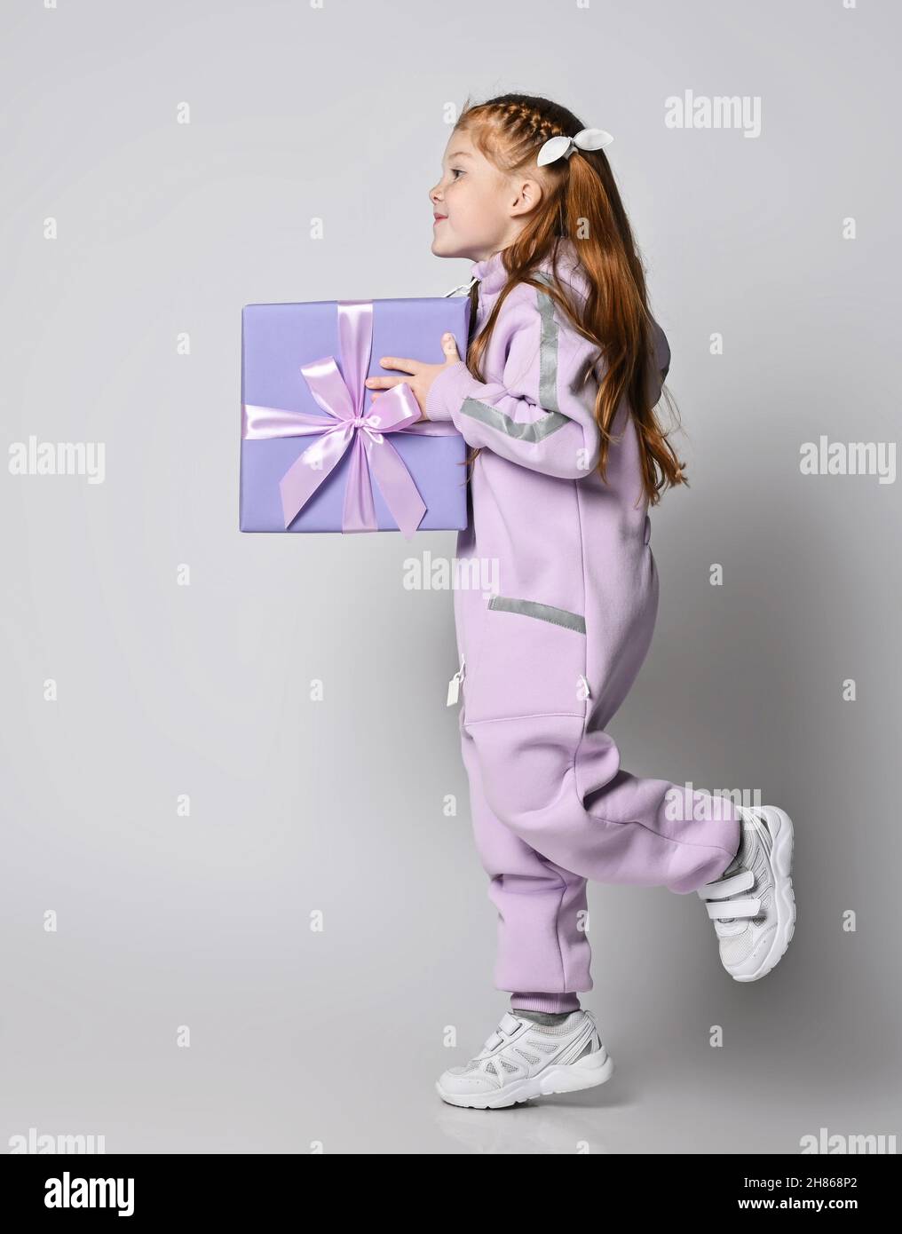 Frolic smiling kid girl in pink jumpsuit is running with big blue gift box with ribbon and bow in hands. Side view Stock Photo