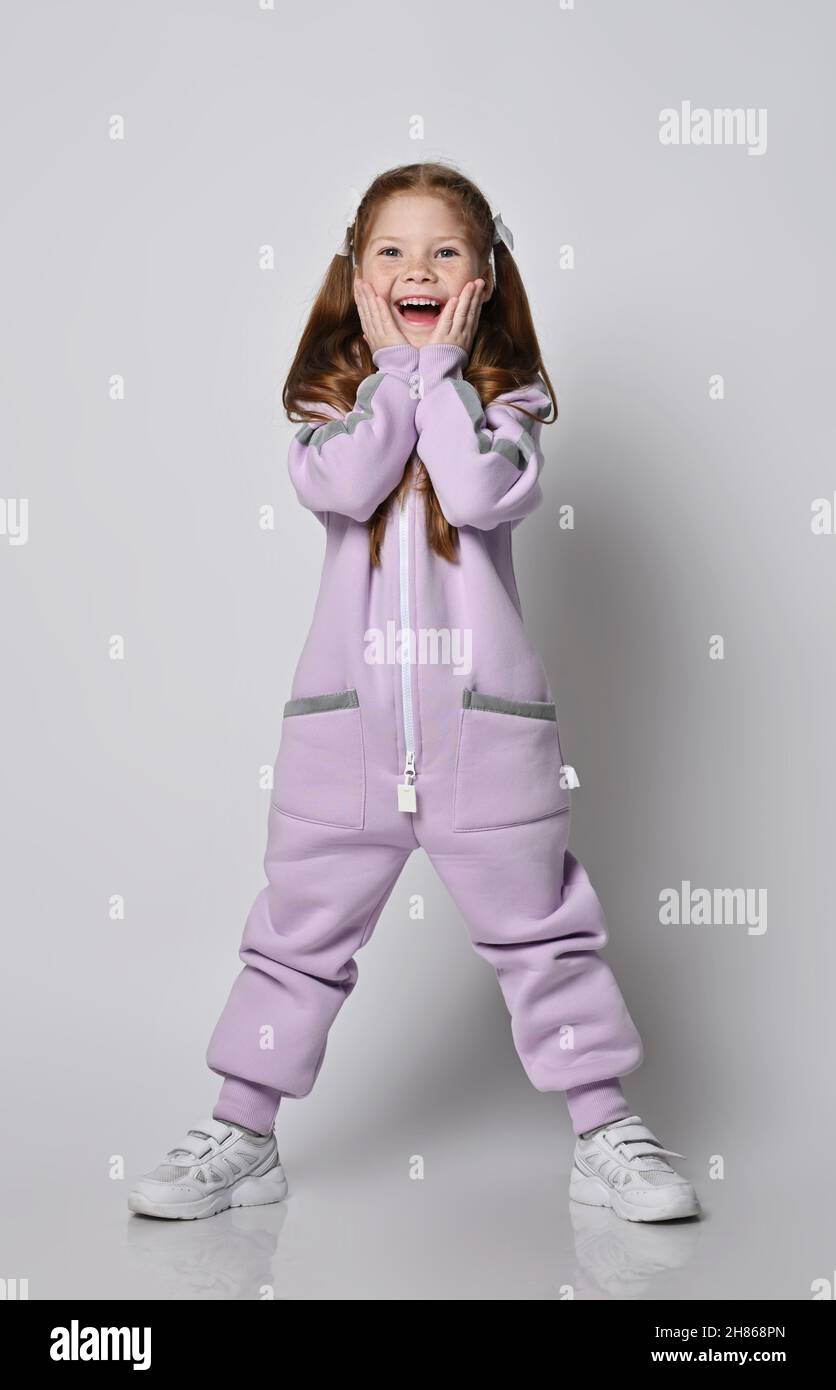 Happy screaming, amazed kid girl in pink jumpsuit with zipper stands holding her mouth open and hands at cheeks Stock Photo