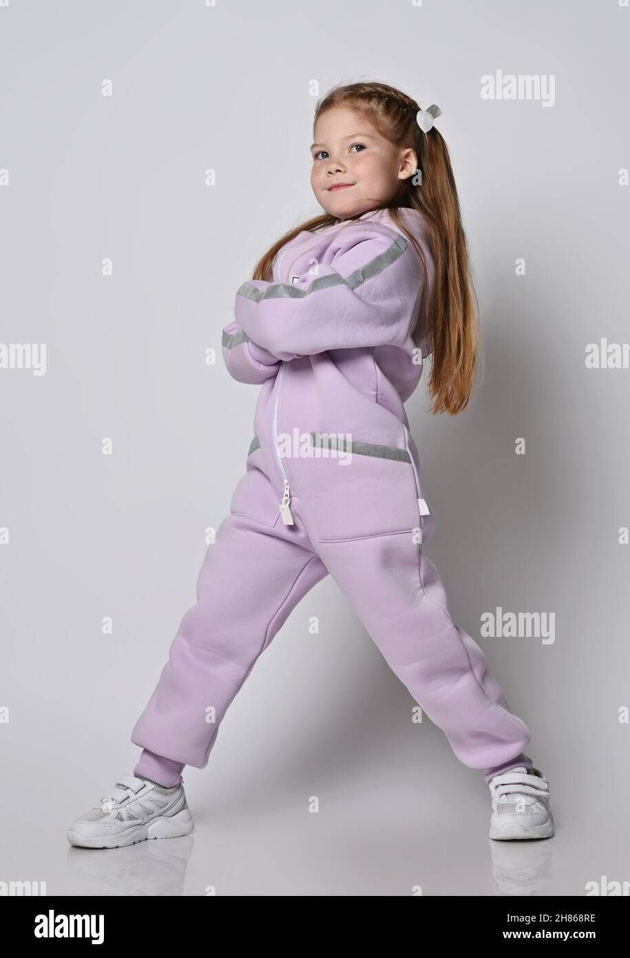 Frolic proudful kid girl in pink modern jumpsuit stands with legs wide apart, arms crossed at chest Stock Photo