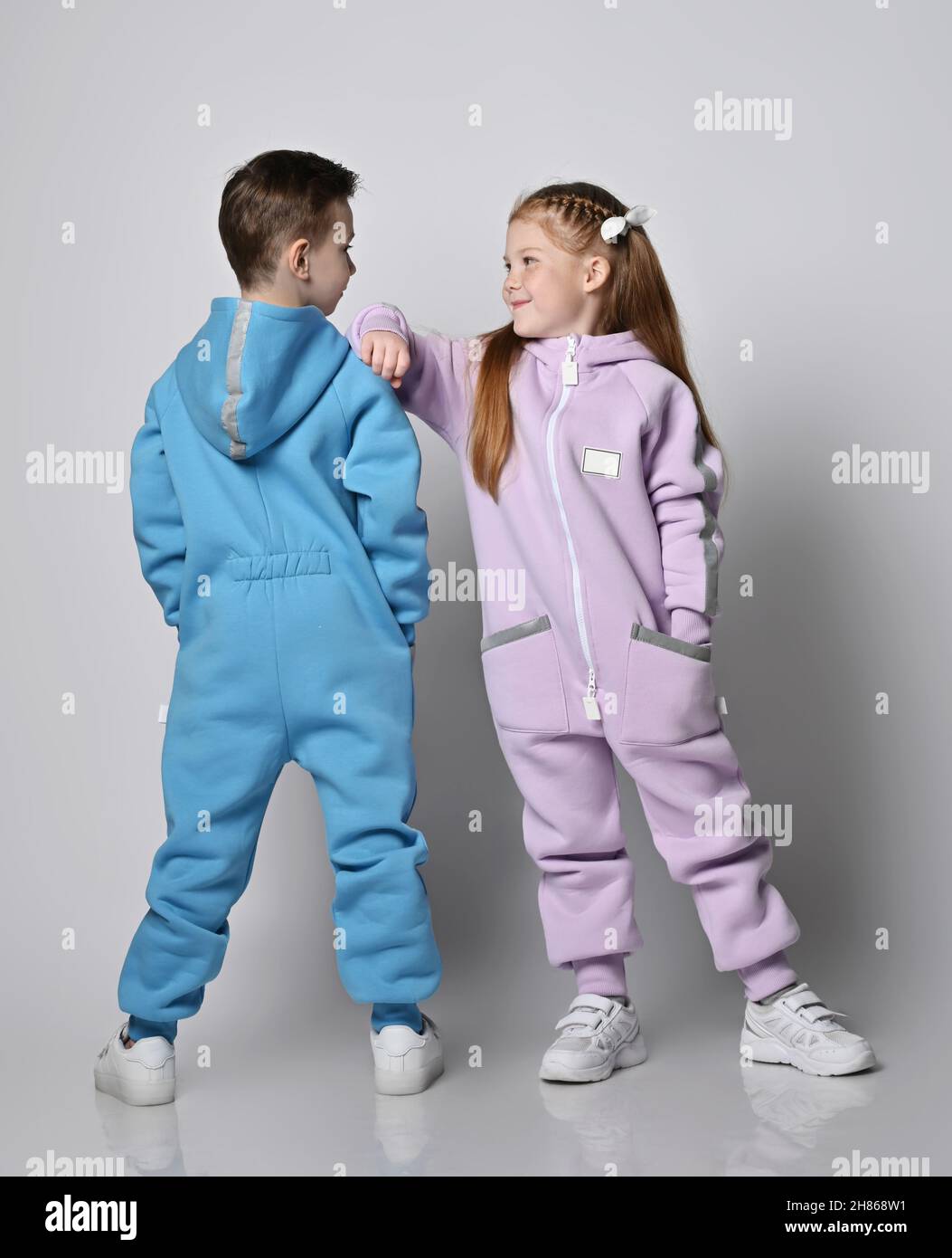 Two cute kids in blue and pink jumpsuits are standing next to each other, boy with his back to us and girl with her face Stock Photo