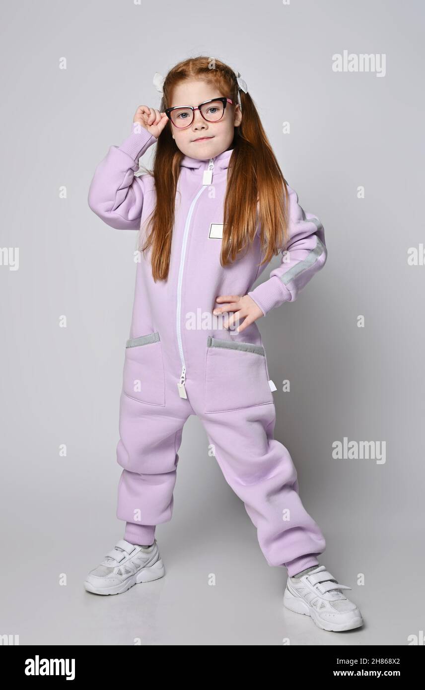 Cool red-haired kid girl in glasses and pink jumpsuit with zipper and big pockets stands with legs wide apart Stock Photo