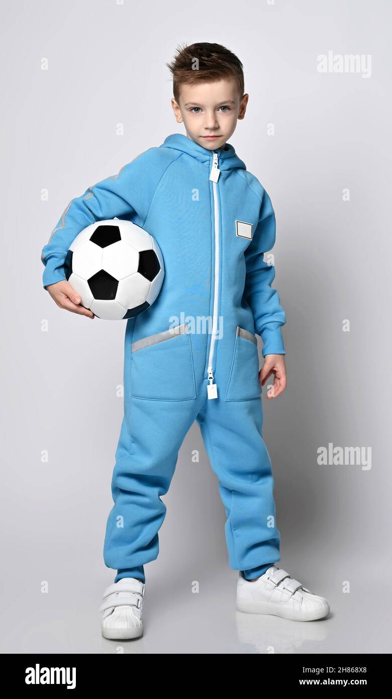 Frolic, active kid boy in blue jumpsuit with hood and pockets with reflective stripes stands holding soccer ball Stock Photo