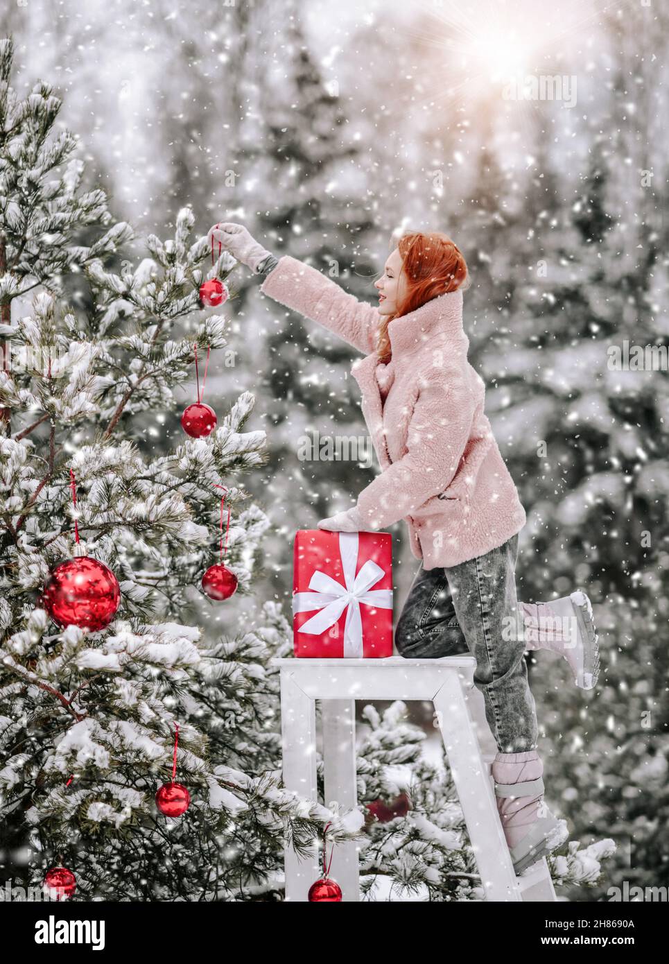 Young woman in warm clothing pink fur coat standing and decorating Christmas tree with balls outdoors in winter snowy forest Stock Photo