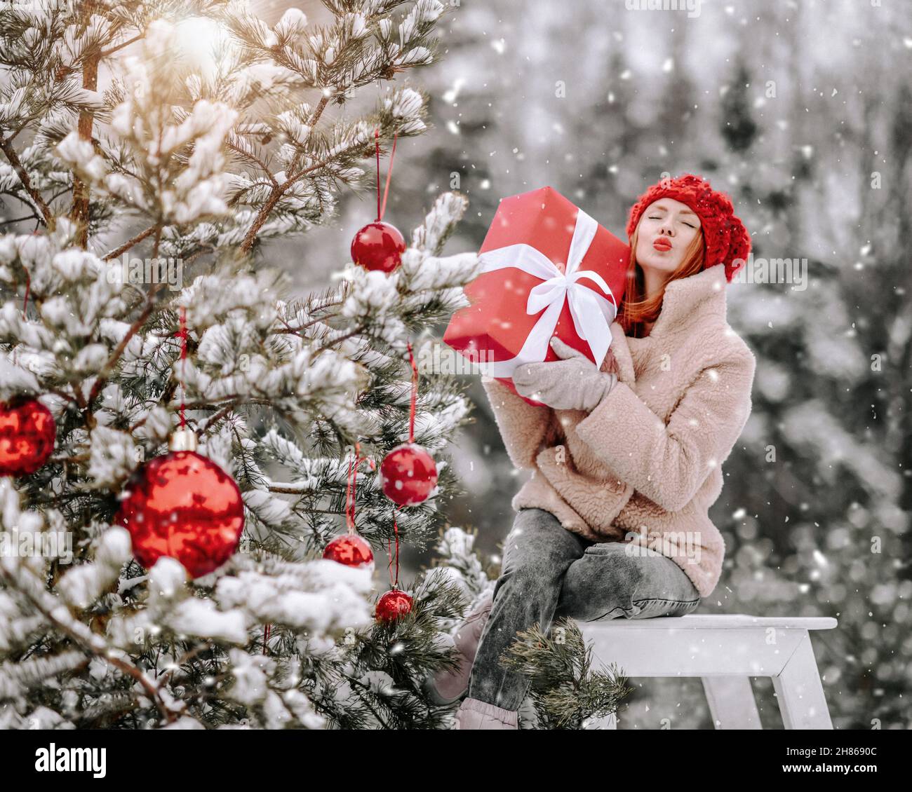 Young positive pretty woman sitting with holiday gift on stool near decorated Christmas outdoors and sending air kiss Stock Photo