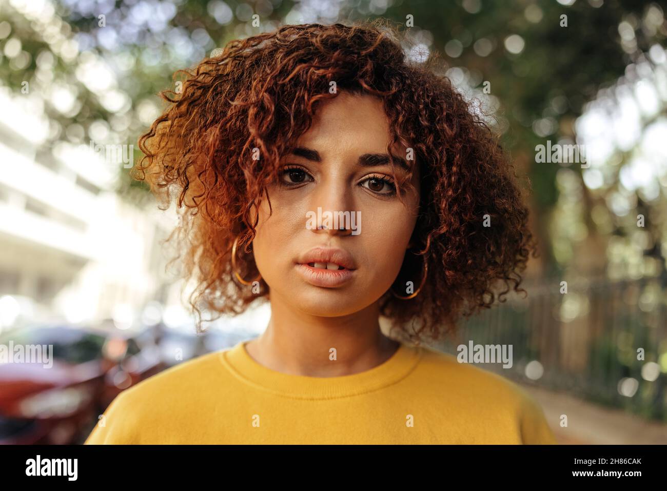 Closeup of a teenage girl looking at the camera. Portrait of a beautiful female youngster wearing red hair and a mustard sweatshirt in the city. Young Stock Photo