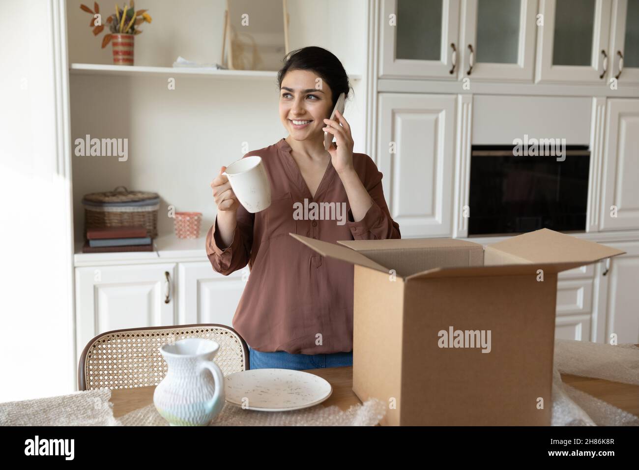 Smiling young indian woman holding phone call conversation, unpacking box. Stock Photo