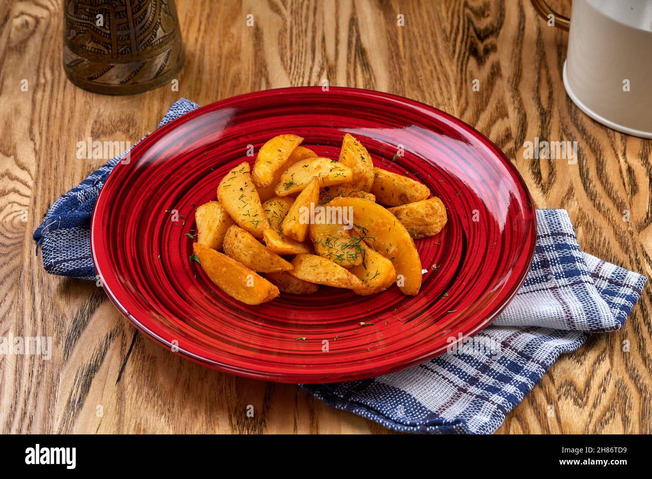 Baked potato wedges with rosemary in red dish, on a blue towel, angle view. Healthy vegan food concept. Stock Photo
