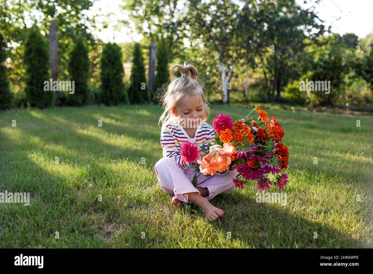 portrait of a beautiful little girl with a bouquet of flowers sitting on a green lawn barefoot. Stock Photo