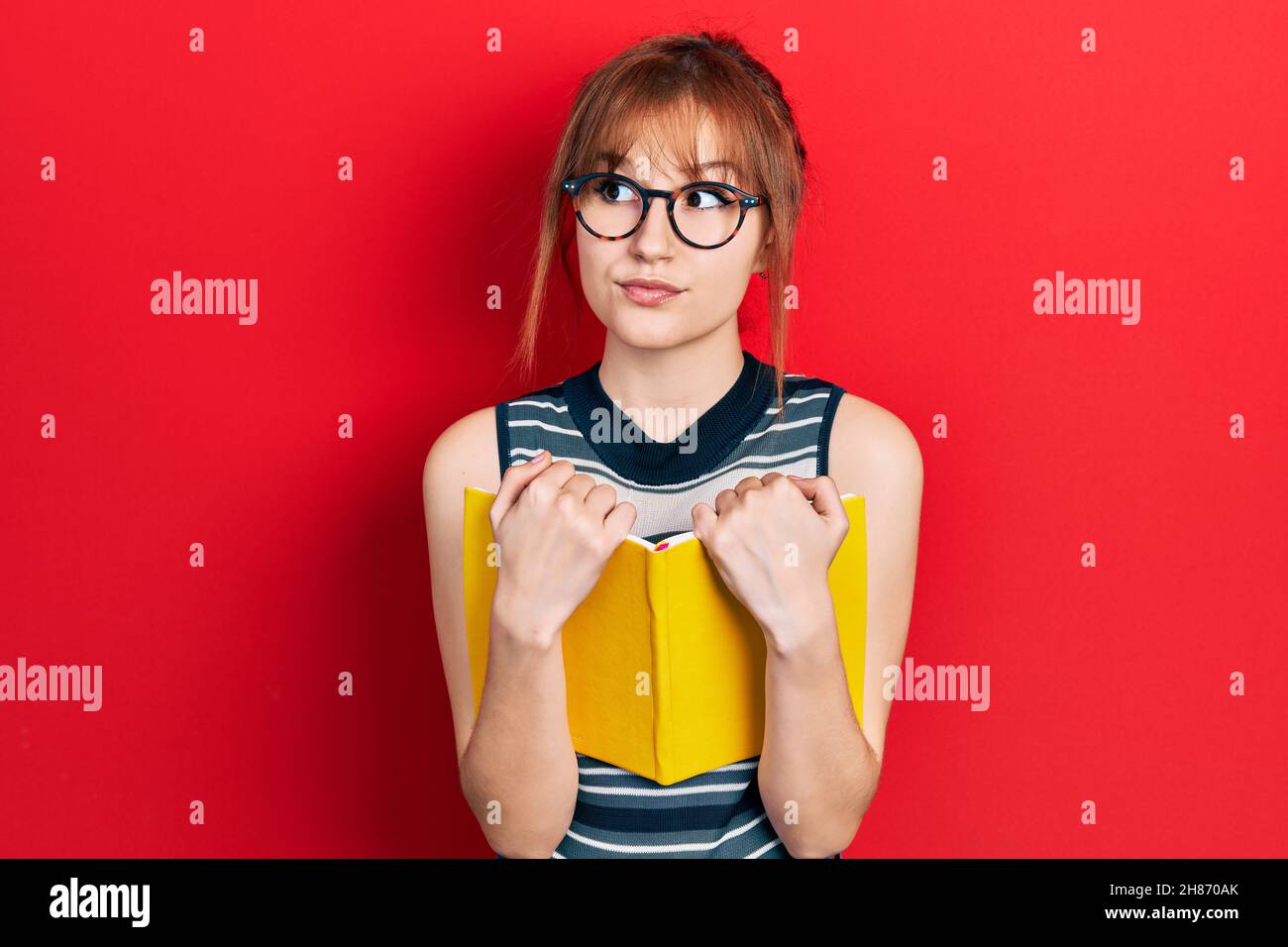 Redhead young woman reading a book wearing glasses smiling looking to
