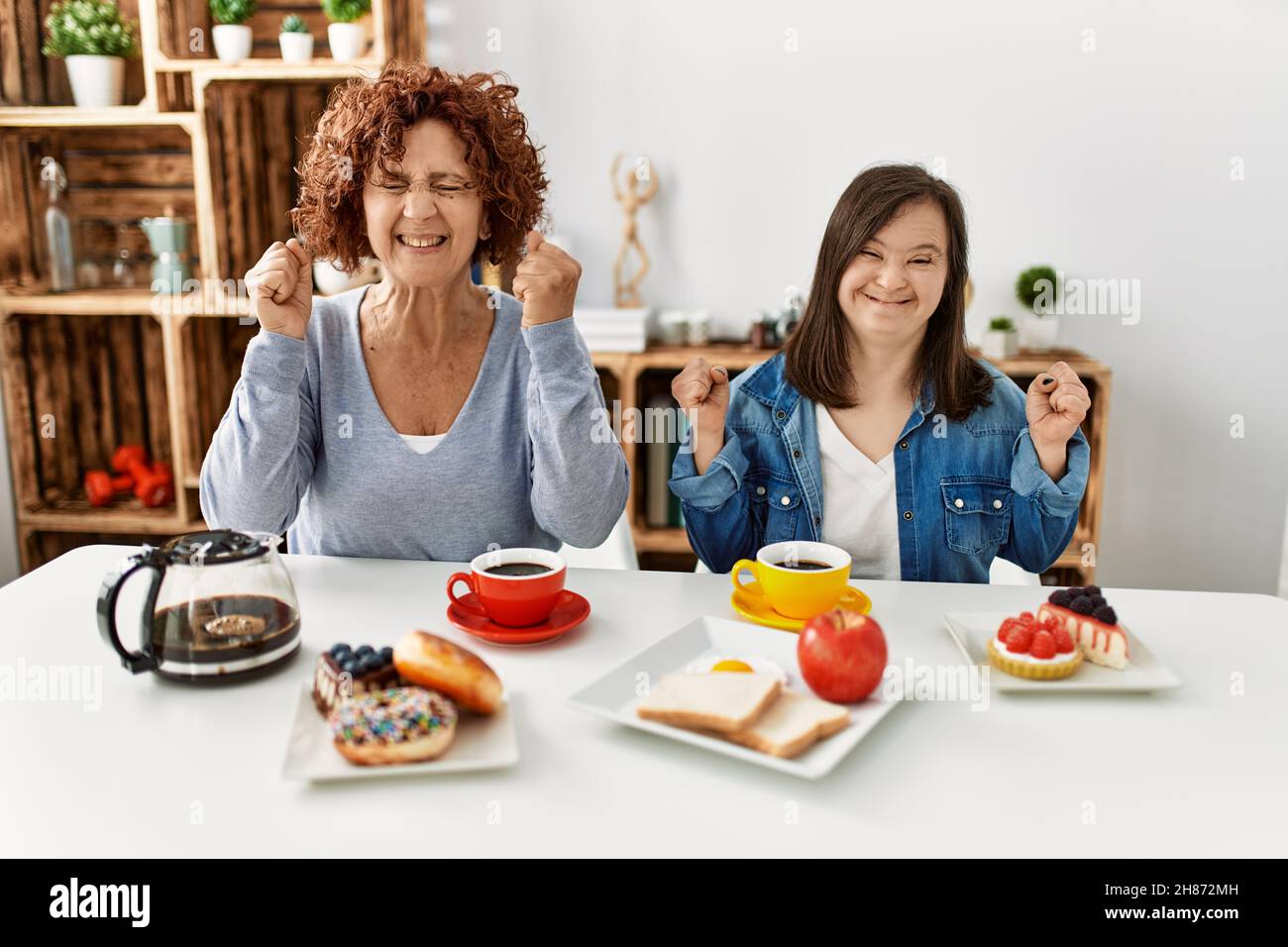 Family of mother and down syndrome daughter sitting at home eating breakfast excited for success with arms raised and eyes closed celebrating victory Stock Photo