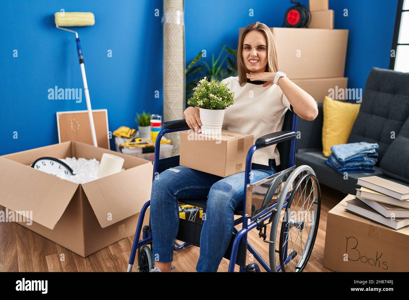 Young woman sitting on wheelchair moving to a new home cutting throat with hand as knife, threaten aggression with furious violence Stock Photo