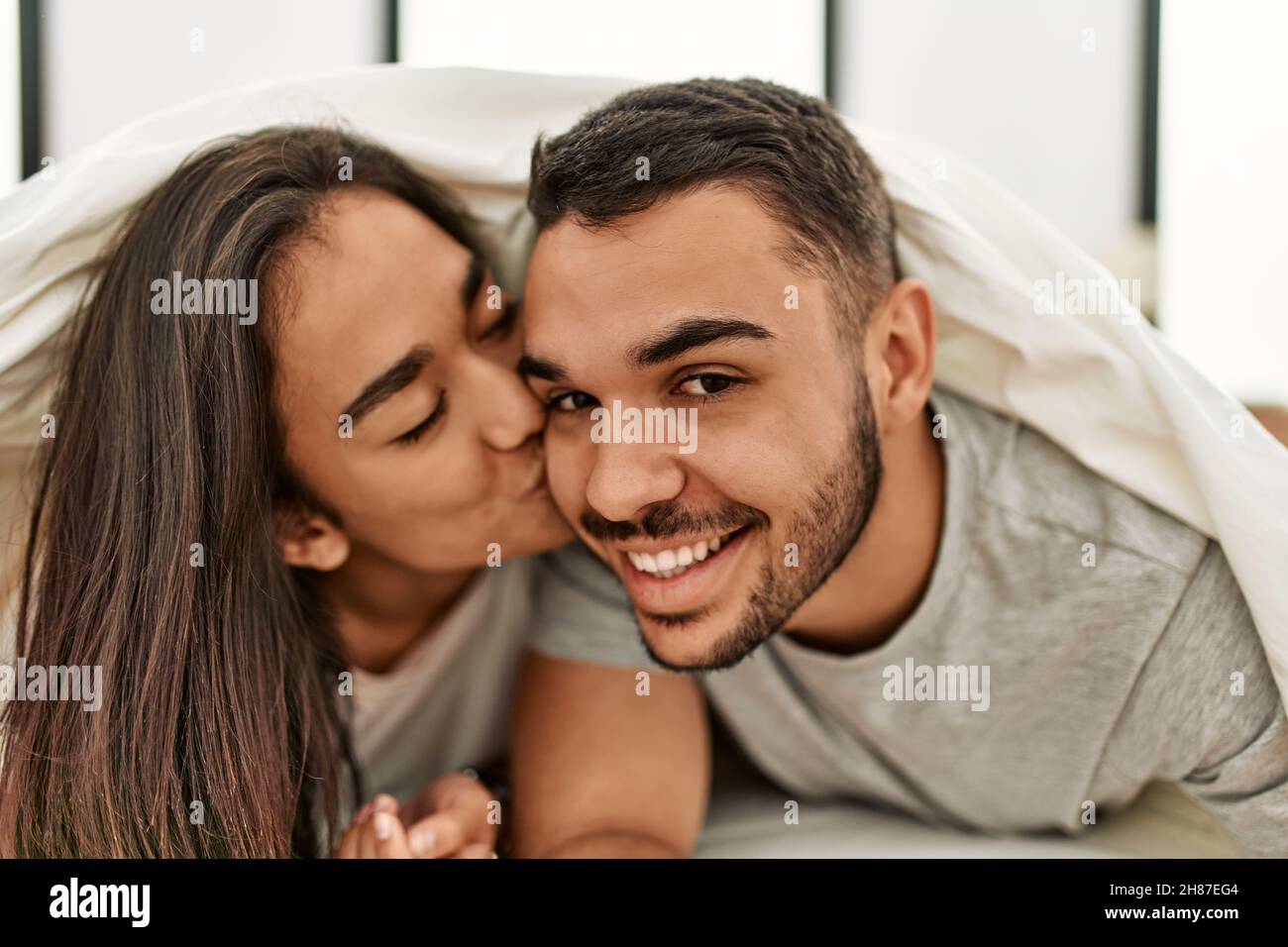 Young latin couple kissing and covering with bed sheet lying on the bed