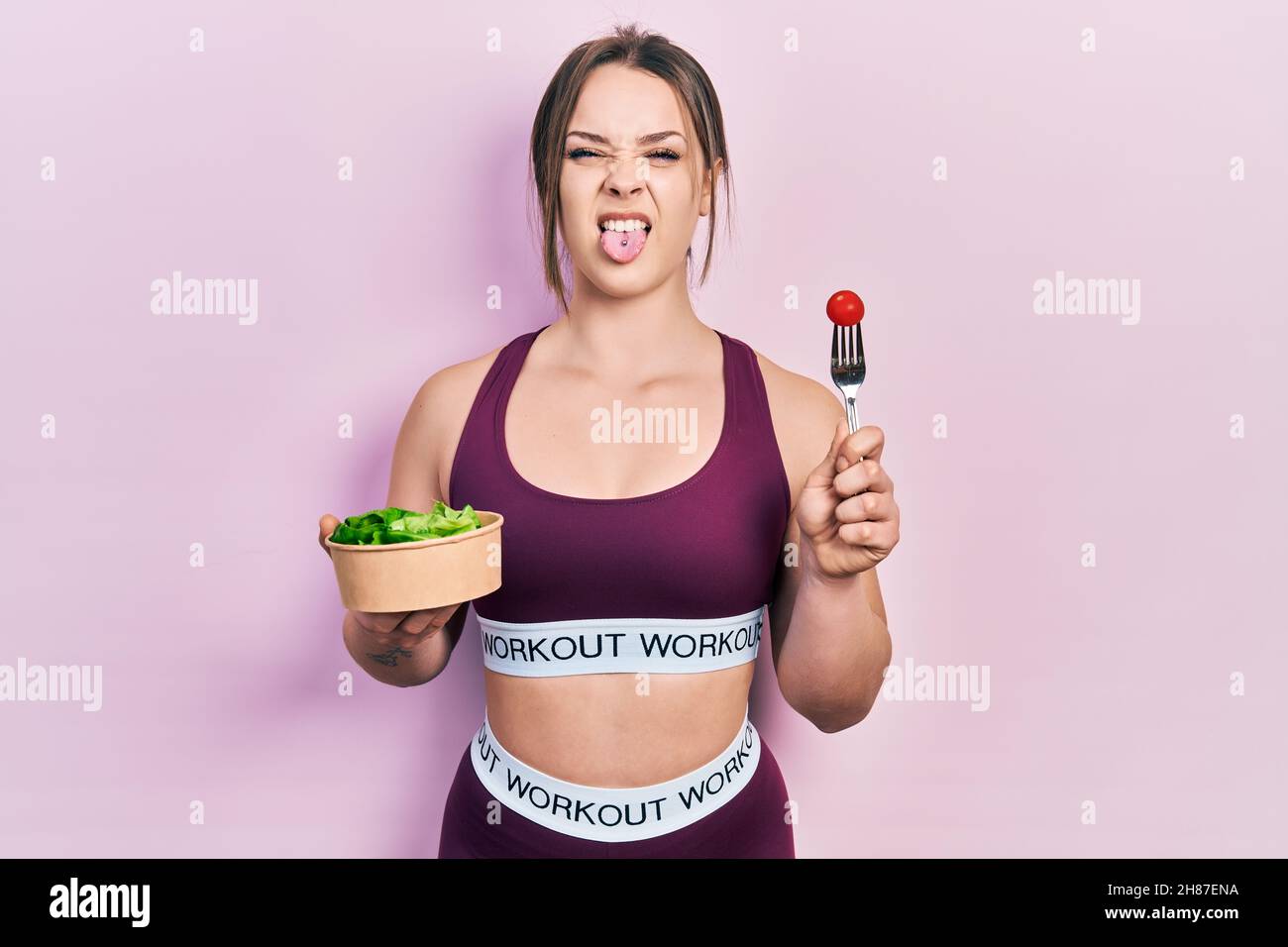 Young hispanic girl wearing sportswear eating salad sticking tongue out happy with funny expression. Stock Photo