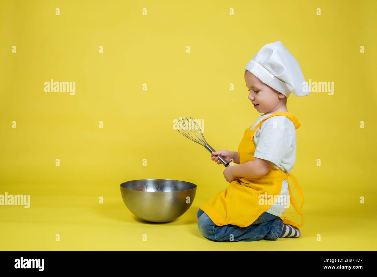Little caucasian Boy playing chef, boy in apron and chef's hat sitting on the floor with a bowl and whisk on yellow background Stock Photo